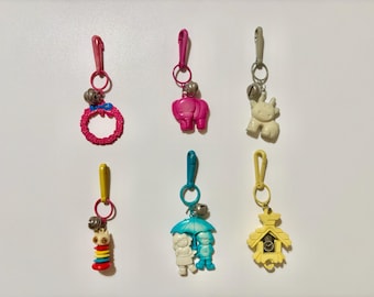 May include: A set of seven colorful keychains with various charms, including a pink elephant, a white elephant, a pink wreath, a blue umbrella with two figures, a yellow cuckoo clock, a colorful stacked ring, and a silver bell.