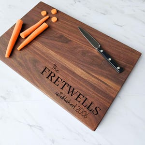 May include: A personalized walnut wood cutting board with the text "The Fretwells established 2006" engraved on the surface. The board is shown with a knife and carrots on top.