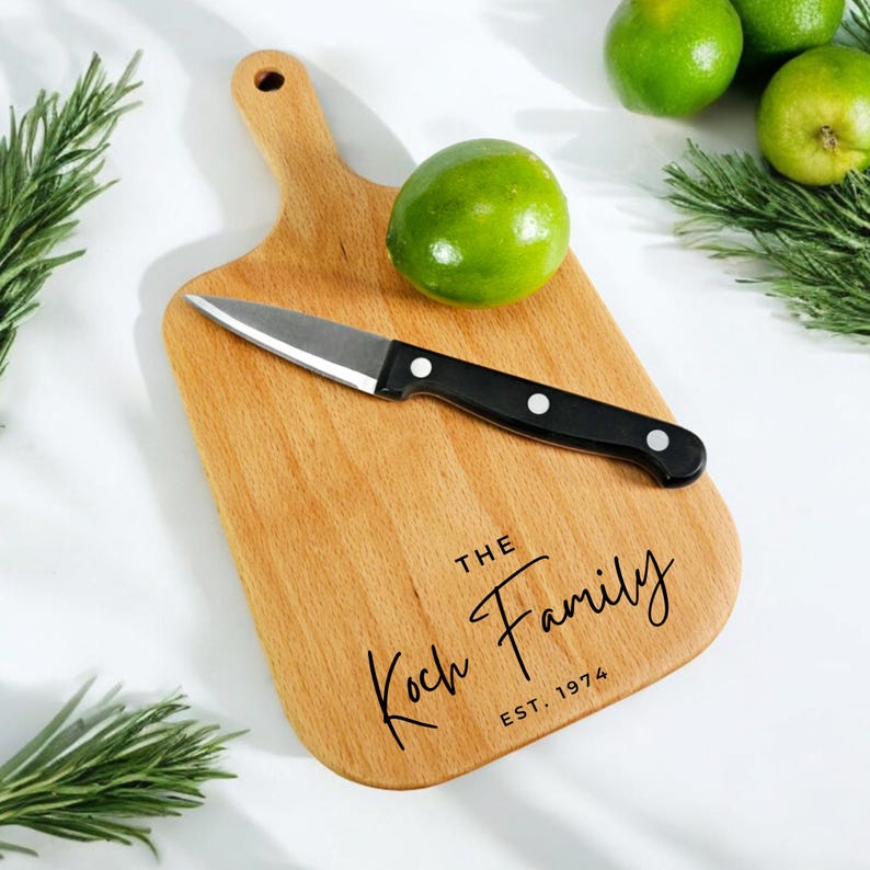 Op de afbeelding: Een houten snijplank met een handvat, een klein mes en een limoen. Op de plank staat "The Koch Family EST. 1974". Het mes heeft een zwart handvat. Groene limoenen en rozemarijntakjes staan op de achtergrond.