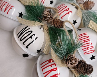 May include: A set of six white jingle bells with red and black lettering. The bells are decorated with burlap ribbon and pine sprigs with pine cones. The words on the bells are "Cheer", "Hope", "Noel", "Believe", "Ho Ho Ho", and "Merry".