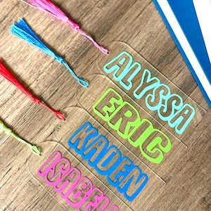 May include: Five clear acrylic bookmarks with colorful names printed on them. The names are Alyssa, Eric, Kaden, and Isabella. Each bookmark has a colorful tassel.