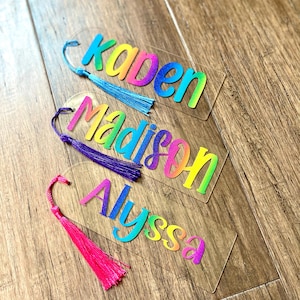 May include: Three clear acrylic bookmarks with personalized names in rainbow gradient colors. Each bookmark has a matching tassel. The names are Kaden, Madison, and Alyssa. The tassels are blue, purple, and pink.