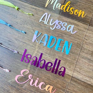 May include: Five clear acrylic bookmarks with different names written in colorful, iridescent lettering. The names are Madison, Alyssa, Kaden, Isabella, and Erica.
