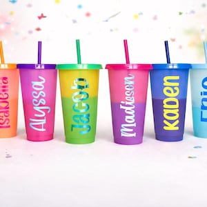 May include: Six colorful, personalized tumblers with lids and straws. Each tumbler is a different color, with names like "Isabella" and "Eric" in a contrasting font. The tumblers are arranged on a white surface with confetti.