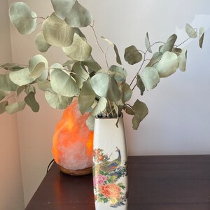 Könnte beinhalten: Eine rechteckige, cremefarbene Keramikvase mit Pfau- und Blumendesign. Die Vase enthält getrocknete Eukalyptuszweige. Die Vase ist etwa 25 cm hoch und steht auf einer dunklen Holzoberfläche.