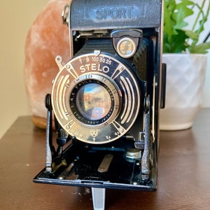 Könnte beinhalten: Eine Vintage-Kamera in Schwarz und Silber mit der Aufschrift "SPORT" und dem Markennamen "STELO" auf dem Objektiv. Das Objektiv hat eine Brennweite von 10,5 cm. Die Kamera ist geöffnet und zeigt das Objektiv und die Innenteile. Die Kamera steht auf einer Holzoberfläche.