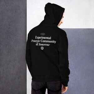 Puede incluir: Sudadera con capucha negra con el texto "Experimental Prototype Community of Tomorrow" y un pequeño gráfico de un globo terráqueo en la espalda.