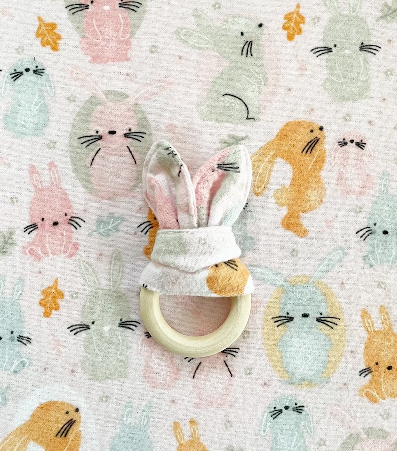 Peut inclure: Un anneau de dentition en forme de lapin rose et blanc avec un anneau en bois. L'anneau de dentition est sur un tissu rose avec un motif de lapins et de feuilles.