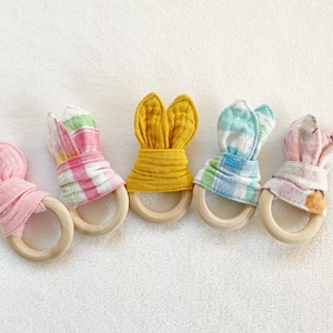 Peut inclure: Cinq anneaux de dentition en bois avec des oreilles de lapin en tissu. Les anneaux de dentition sont en rose, jaune, bleu et en vichy rose et blanc.