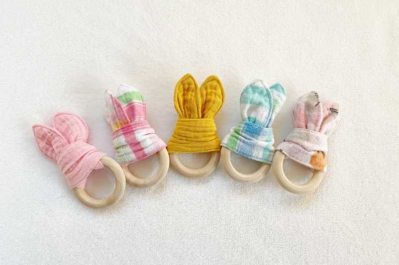Peut inclure: Cinq anneaux de dentition en bois avec des oreilles de lapin en tissu. Les anneaux de dentition sont en rose, jaune, bleu et en vichy rose et blanc.