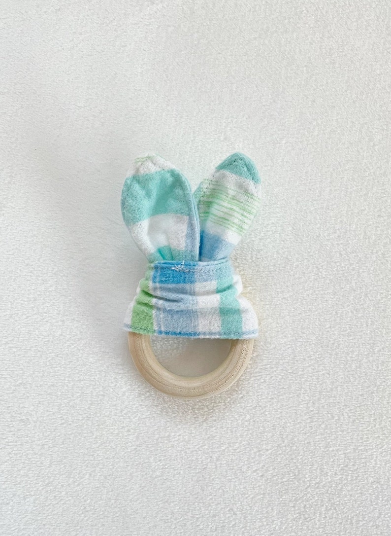 Peut inclure: Un anneau de dentition en bois avec un tissu &agrave; carreaux bleus, verts et blancs nou&eacute; autour, formant des oreilles de lapin.