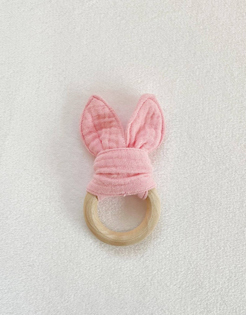 Peut inclure: Hochet en tissu rose en forme de lapin avec un anneau en bois. Le tissu est nou&eacute; au centre de l'anneau.