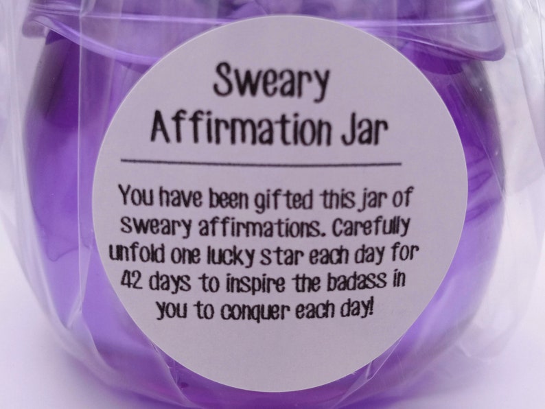 May include: A purple glass jar wrapped in clear plastic, featuring a white circular label with black text that reads: Sweary Affirmation Jar. You have been gifted this jar of sweary affirmations. Carefully unfold one lucky star each day for 42 days to inspire the badass in you to conquer each day!