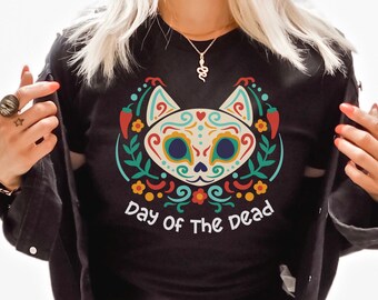 May include: A black t-shirt featuring a colorful graphic of a cat face designed with intricate patterns in red, yellow, teal, and white. The cat is surrounded by floral and chili pepper motifs. Below the graphic, the text reads Day Of The Dead.