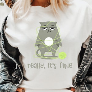 May include: White t-shirt featuring a grumpy gray cat tangled in neon green yarn, with a matching ball of yarn. The text "really, it's fine" is printed below the cat. The shirt is a casual, everyday wear item.
