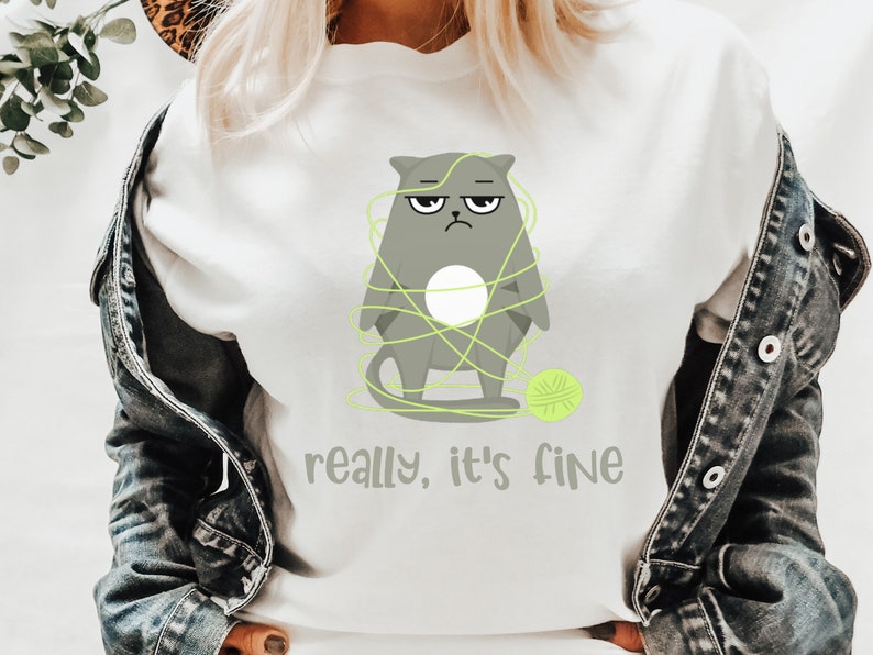 May include: White t-shirt featuring a grumpy gray cat tangled in neon green yarn, with a matching ball of yarn. The text "really, it's fine" is printed below the cat. The shirt is a casual, everyday wear item.