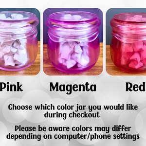 May include: Three glass jars with screw-top lids, each filled with small folded paper stars. The jars are tinted pink, magenta, and red. Text below reads: Pink, Magenta, Red. Choose which color jar you would like during checkout. Please be aware colors may differ depending on computer/phone settings.