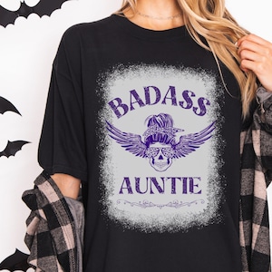 May include: Black t-shirt with a purple graphic that reads "BADASS AUNTIE". The graphic features a skull wearing sunglasses and a bandana, with wings. The shirt is a casual style.