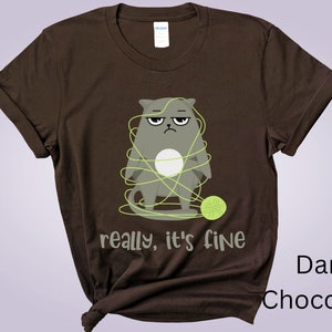 May include: Dark chocolate brown t-shirt featuring a grumpy gray cat tangled in green yarn. The text "really, it's fine" is below the cat. A green ball of yarn is on the right. The shirt is a Gildan brand.