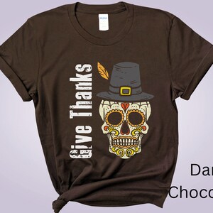 May include: A dark chocolate brown short-sleeve t-shirt featuring a graphic of a skull wearing a pilgrim hat with a feather. The text "Give Thanks" is printed vertically in white next to the skull, which has orange and yellow decorative patterns.