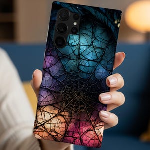 May include: A rectangular smartphone case featuring a vibrant, multi-colored spider web pattern. The design transitions from deep blue and purple at the top to warm orange and pink tones at the bottom, with intricate black web lines throughout.