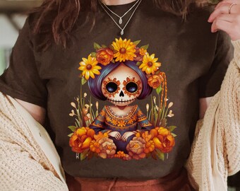 May include: A dark brown t-shirt featuring a graphic illustration of a stylized character with a skull face, dark hair, and a floral crown. The character wears a patterned dress and is surrounded by vibrant orange and yellow flowers and tall grass.