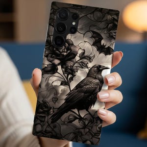 May include: A rectangular smartphone case featuring a detailed graphic print of a black raven perched among dark roses and flying bats. The design uses a monochromatic color palette with shades of black, gray, and cream, showcasing a complex pattern.