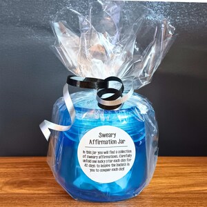 May include: A translucent blue plastic jar wrapped in clear cellophane with black and white curling ribbons. A white circular label on the front reads: Sweary Affirmation Jar. In this jar you will find a collection of sweary affirmations.