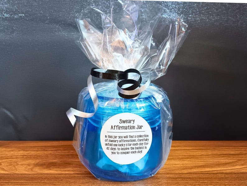 May include: A translucent blue plastic jar wrapped in clear cellophane with black and white curling ribbons. A white circular label on the front reads: Sweary Affirmation Jar. In this jar you will find a collection of sweary affirmations.