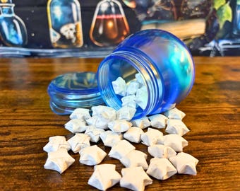 May include: A translucent blue jar with a matching lid lying on its side, spilling out a collection of small, white, folded paper stars. The jar has a rounded shape and a smooth, glossy finish, contrasting with the matte texture of the paper stars.