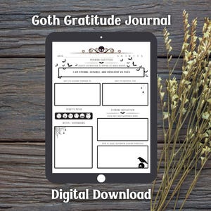 May include: A digital download of a Goth Gratitude Journal displayed on a tablet. The journal features a skull and bat design, with prompts for daily reflection. The words "I AM STRONG, CAPABLE, AND RESILIENT AS FUCK" are visible. The background is a wooden surface.