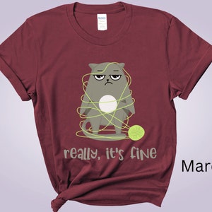 May include: Maroon t-shirt featuring a grumpy cartoon cat tangled in yellow yarn, with a matching ball of yarn at its feet. The text "really, it's fine" is printed below the cat. The shirt is made of a soft material.
