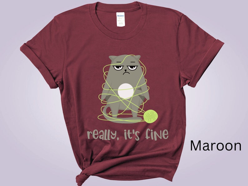 May include: Maroon t-shirt featuring a grumpy cartoon cat tangled in yellow yarn, with a matching ball of yarn at its feet. The text "really, it's fine" is printed below the cat. The shirt is made of a soft material.