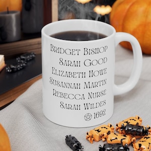 May include: A white ceramic mug featuring black printed text listing the names Bridget Bishop, Sarah Good, Elizabeth Howe, Susannah Martin, Rebecca Nurse, and Sarah Wildes, followed by a pentagram symbol and the year 1692 in a serif font.