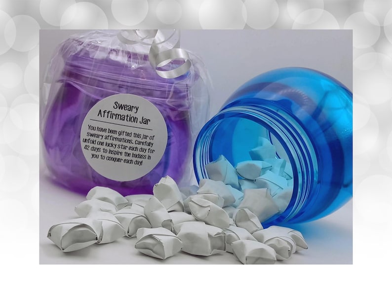 May include: A purple plastic jar with a white label reading Sweary Affirmation Jar and a blue plastic jar tipped on its side, spilling out numerous small, white, folded paper stars. The purple jar is wrapped in clear plastic with a white ribbon.