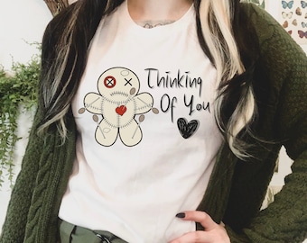 May include: A white t-shirt featuring a graphic of a beige voodoo doll with a red heart, stitched details, and pins. The text "Thinking Of You" is printed in black script next to a hand-drawn black heart icon on the front of the garment.