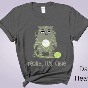 May include: Dark heather gray t-shirt featuring a grumpy cartoon cat tangled in yarn with a ball of yarn. The text "really, it's fine" is printed below the cat. The shirt is a Gildan brand.