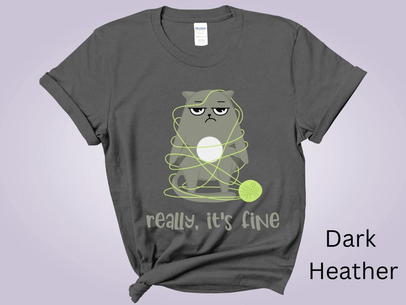 May include: Dark heather gray t-shirt featuring a grumpy cartoon cat tangled in yarn with a ball of yarn. The text "really, it's fine" is printed below the cat. The shirt is a Gildan brand.