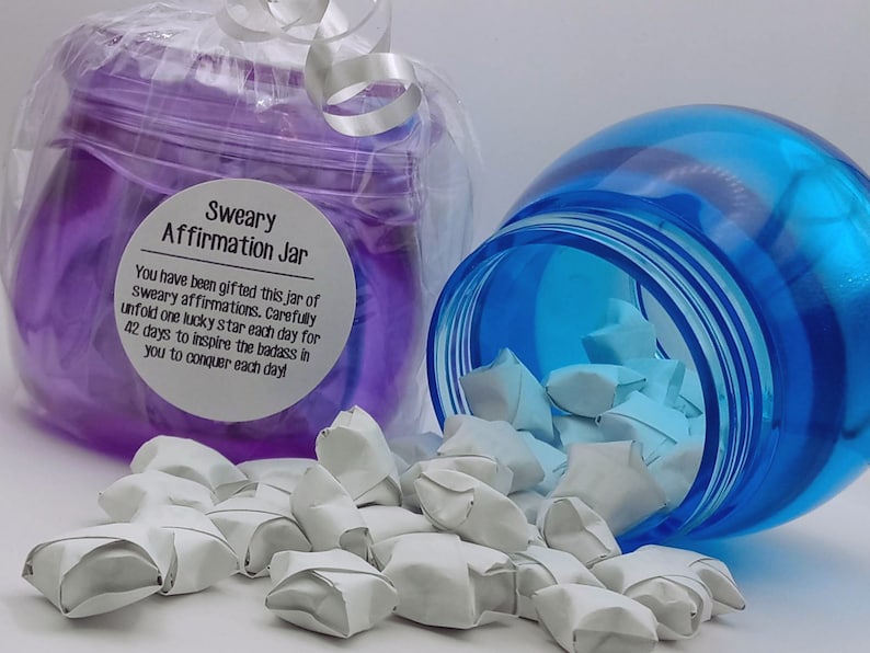May include: A purple jar wrapped in clear plastic with a white label reading Sweary Affirmation Jar and a blue jar tipped on its side, both filled with small white paper stars. The label text includes instructions to unfold one star each day for 42 days.