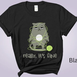 May include: Black t-shirt featuring a grumpy gray cat tangled in green yarn, with a matching ball of yarn. The text "really, it's fine" is printed below the cat. The shirt is made of a soft material.