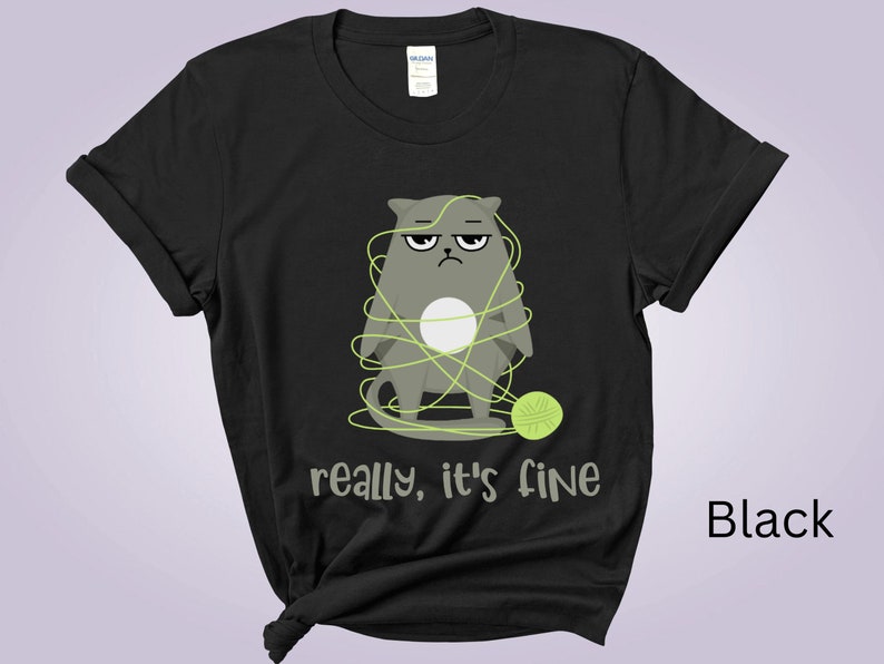 May include: Black t-shirt featuring a grumpy gray cat tangled in green yarn, with a matching ball of yarn. The text "really, it's fine" is printed below the cat. The shirt is made of a soft material.