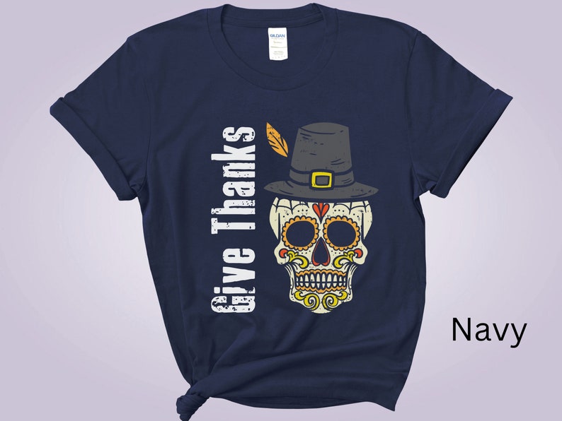 May include: A navy blue short-sleeve t-shirt featuring a graphic of a sugar skull wearing a pilgrim hat with a feather. The text "Give Thanks" is printed vertically in white next to the skull. The skull has yellow, orange, and red details.