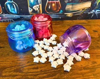 May include: Three small, round, translucent glass jars in blue, red, and purple, each with a threaded rim. The blue and red jars are upright and filled with small, folded paper stars, while the purple jar is tipped over, spilling white paper stars.