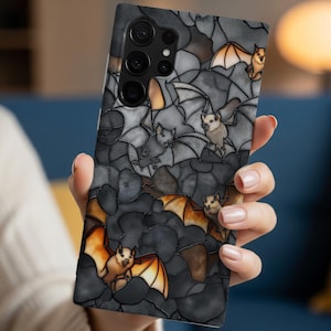 May include: A phone case featuring a stained-glass design with bats. The bats have orange and white wings, set against a dark gray background. The case has a glossy finish and a multi-lens camera.