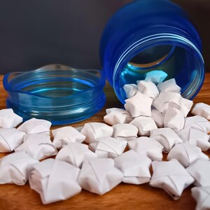 May include: A translucent blue plastic jar with a matching lid lying on its side, spilling out numerous small, white, five-pointed paper stars folded into three-dimensional shapes. The jar has a wide mouth and a threaded rim for the lid.