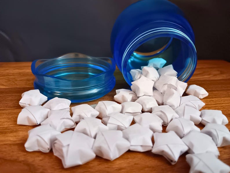 May include: A translucent blue plastic jar with a matching lid lying on its side, spilling out numerous small, white, five-pointed paper stars folded into three-dimensional shapes. The jar has a wide mouth and a threaded rim for the lid.