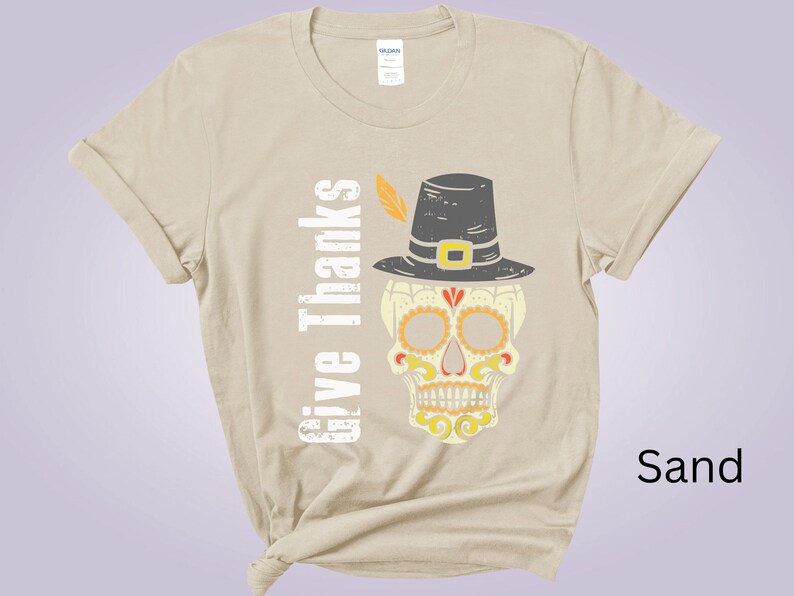 May include: A sand-colored t-shirt featuring a graphic of a skull wearing a pilgrim hat with a feather. The text "Give Thanks" is printed vertically in white next to the skull, which is decorated with yellow and orange patterns.