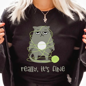 May include: Black crew neck sweatshirt featuring a grumpy gray cat tangled in green yarn with a ball of yarn. The text "really, it's fine" is printed below the cat. The sweatshirt is a casual, comfortable garment.