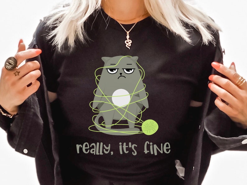 May include: Black crew neck sweatshirt featuring a grumpy gray cat tangled in green yarn with a ball of yarn. The text "really, it's fine" is printed below the cat. The sweatshirt is a casual, comfortable garment.