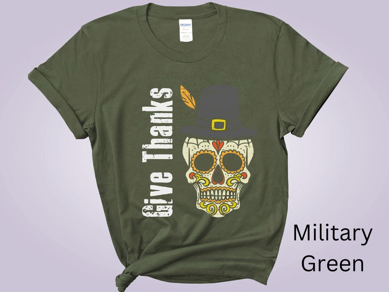 May include: A military green t-shirt featuring a graphic of a sugar skull wearing a pilgrim hat with a feather. The text "Give Thanks" is printed vertically in white to the left of the skull. The skull has orange and yellow decorative patterns.
